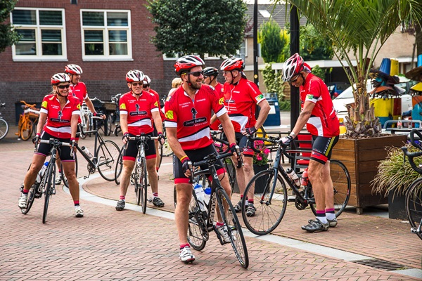 Eine Gruppe von Radfahrern in roter Sportkleidung steht mit ihren Fahrrädern auf einem Platz im Zentrum von Nederweert bereit.