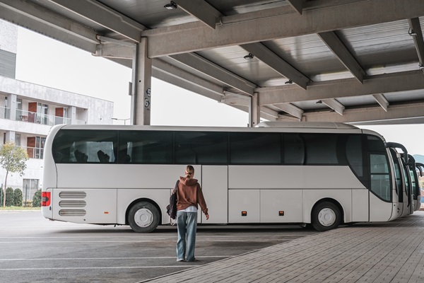 Vrouw met rugzak staat op een overdekte parkeerplaats en kijkt naar een geparkeerde touringcar. In de achtergrond zijn meer bussen en een modern gebouw zichtbaar.