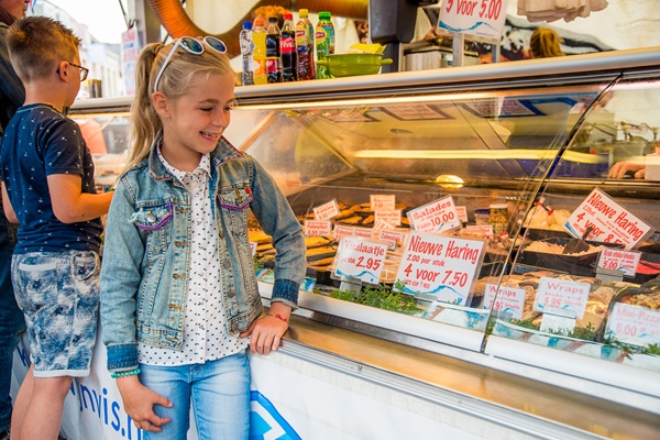 Een glimlachend meisje bij een viskraam op een markt in Weert, met haring en andere verse visproducten in de vitrine.