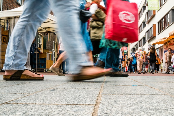 Belebte Einkaufsstraße an einem verkaufsoffenen Sonntag in Weert. Im Vordergrund gehen Menschen vorbei, die sich auf ihre Füße und Taschen konzentrieren. Im Hintergrund sind Einkäufer, Marktstände und Geschäfte zu sehen.